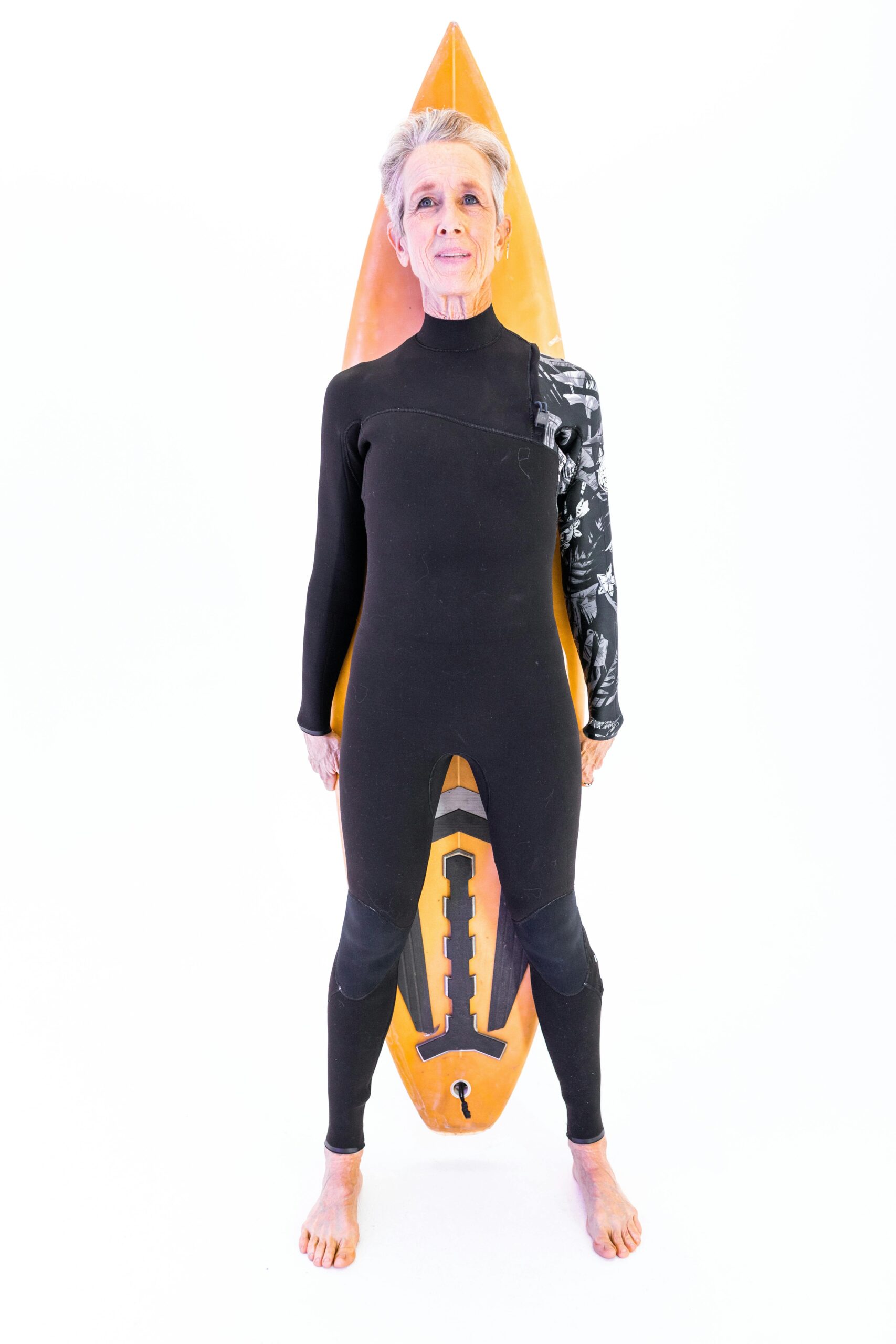 Elderly woman confidently poses in wetsuit holding a surfboard indoors.